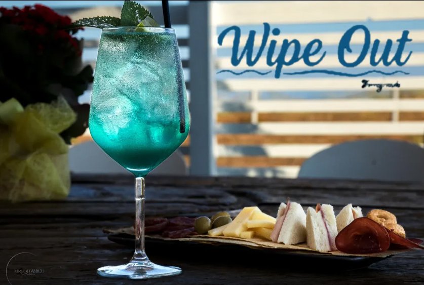 Cocktail azzurro con ghiaccio e menta accanto a tagliere di salumi, formaggio, olive e tramezzini, logo Wipe Out